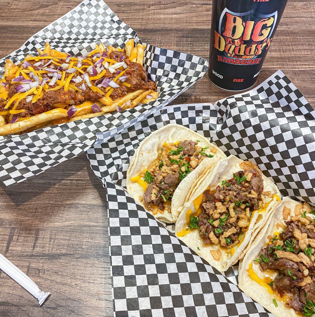 3 prime rib street tacos served on black and white checked paper along side brisket chili cheese fries on black and white checkered paper with a Big Daddy's cup in the back.